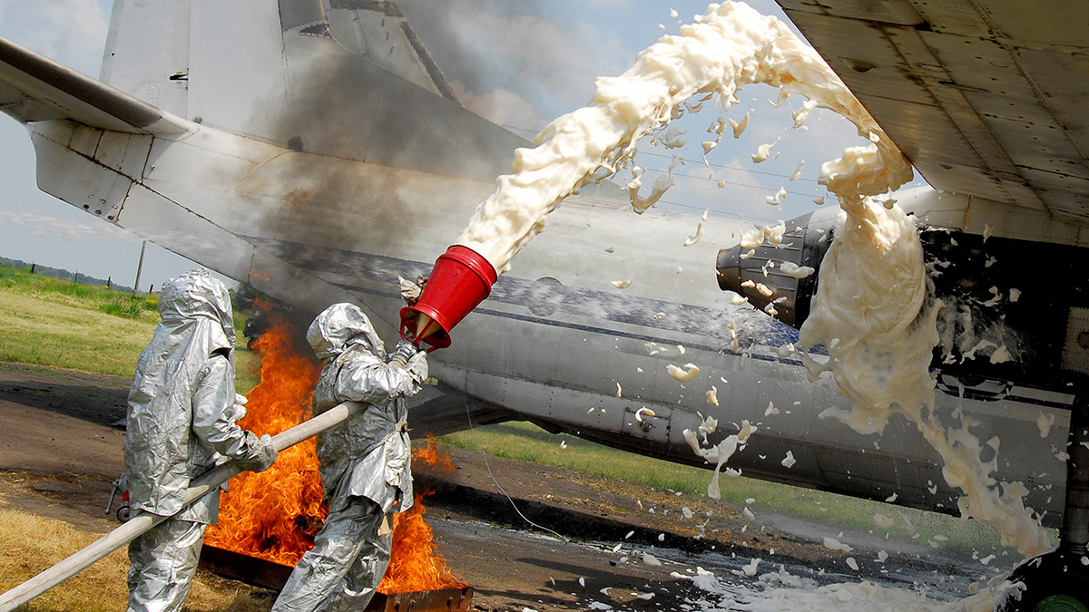 Firefighters in proximity suits applying AFFF aqueous film-forming foam to an aircraft fire during training — PFAS-containing foam applied to aircraft aprons and fire training areas is a primary source of stormwater contamination at military installations and airports